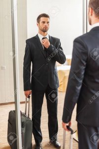 Ready for business trip. Confident young man in formalwear adjusting his necktie while standing against mirror in hotel room